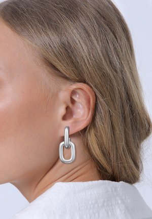 Silberne Metallohrringe mit einem länglichen ovalen Hoop-Design und einem rechteckigen Verbindungsanhänger. Glatte Textur, modernes Format, minimalistische Stil.