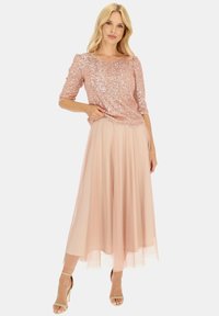 Różowa bluzka z cekinami i rękawami o długości trzech ćwierć, zestawiona z lekką, zwiewną, różową spódnicą maxi. Strój charakteryzuje się warstwowym, teksturowanym wzorem.