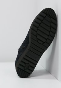 Suela de zapato negra con profundas estrías horizontales y un patrón de dibujo ovalado, situada en ángulo contra una pared blanca y la esquina del suelo.