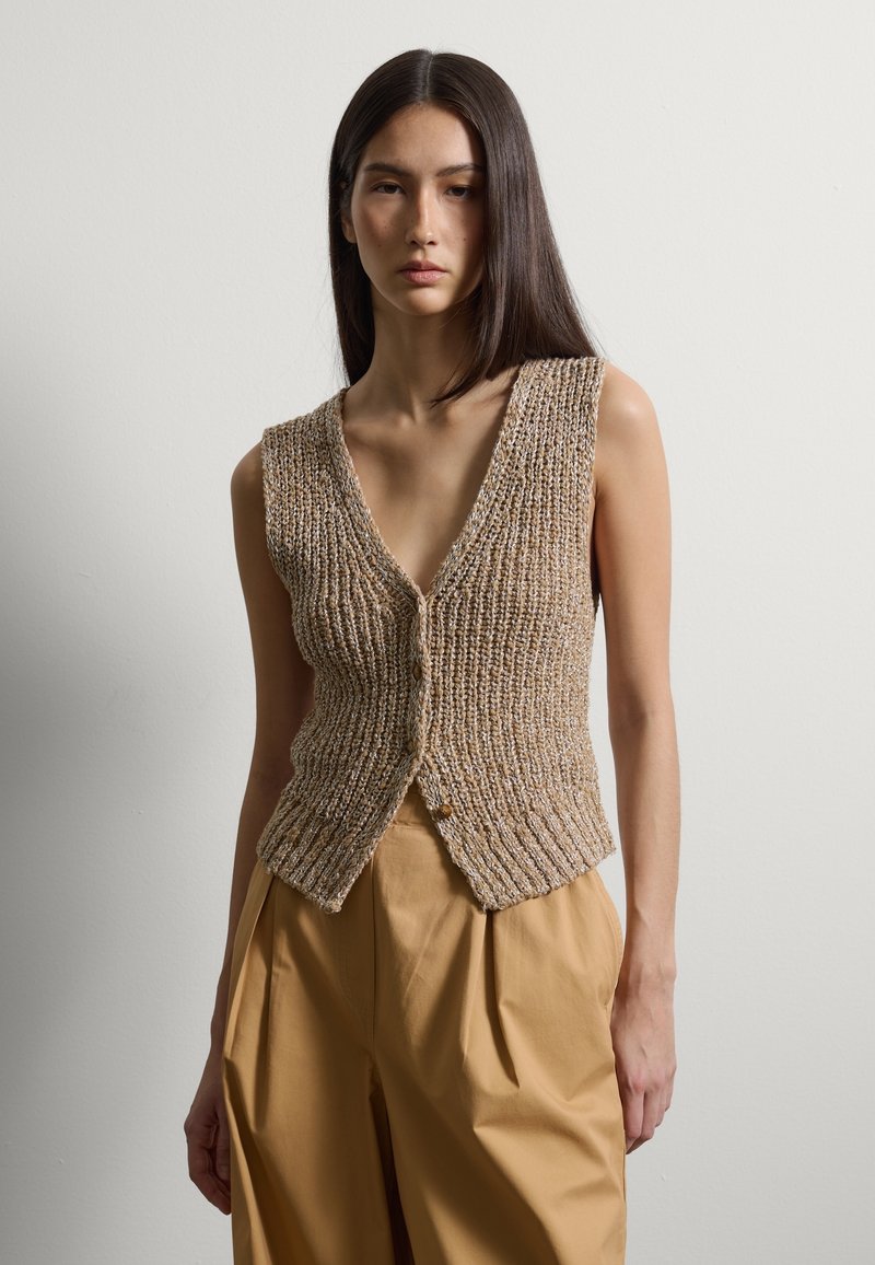 Frau mit glattem, dunklem Haar, die eine strukturierte, ärmellose beige Strickweste und hoch taillierte, weit geschnittene hellbraune Hosen vor einem schlichten Hintergrund trägt.