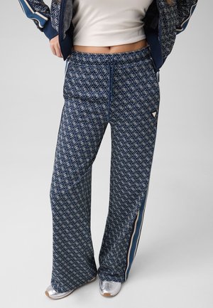 Femme portant un pantalon ample à motifs bleu marine avec bandes latérales, un top court blanc et des baskets argentées, debout devant un fond uni.