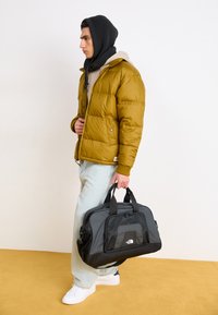 Black and gray duffel bag with mesh accents and logo, carried by a person in a yellow puffer jacket and light jeans on a yellow floor.