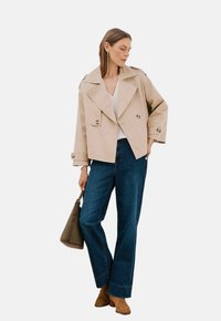 Beige kort trenchcoat med stora knappar, bärs över en vit topp och mörkblå vida jeans. Bruna ankelstövlar och en grön handväska.