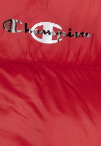 Czerwony materiał wyściełany, z błyszczącym, srebrnym logo "Champion", z wyraźnym owalnym akcentem. Gładka faktura z widocznymi detalami szwów.