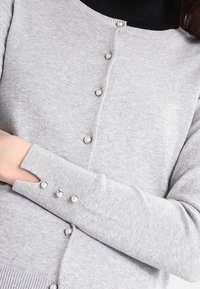Ljusgrå cardigan i mjukt material med rund halsringning, pärl-liknande knappar och utskurna ärmslut med liknande knapppdetaljer.