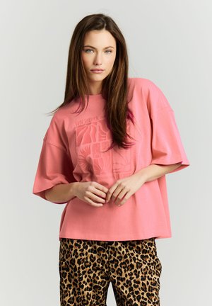 Femme aux longs cheveux bruns portant un t-shirt imprimé rose ample et un pantalon à motif léopard, debout les mains jointes devant.
