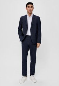 Completo blu navy scuro con giacca a due bottoni, risvolti stretti e pantaloni coordinati. Camicia bianca con colletto sotto. Sneakers bianche completano l'outfit.