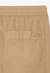 Pantalones cortos beige con cinturilla elástica, que cuentan con un bolsillo trasero y un logo bordado en el lateral. Textura de tela suave y diseño sencillo.