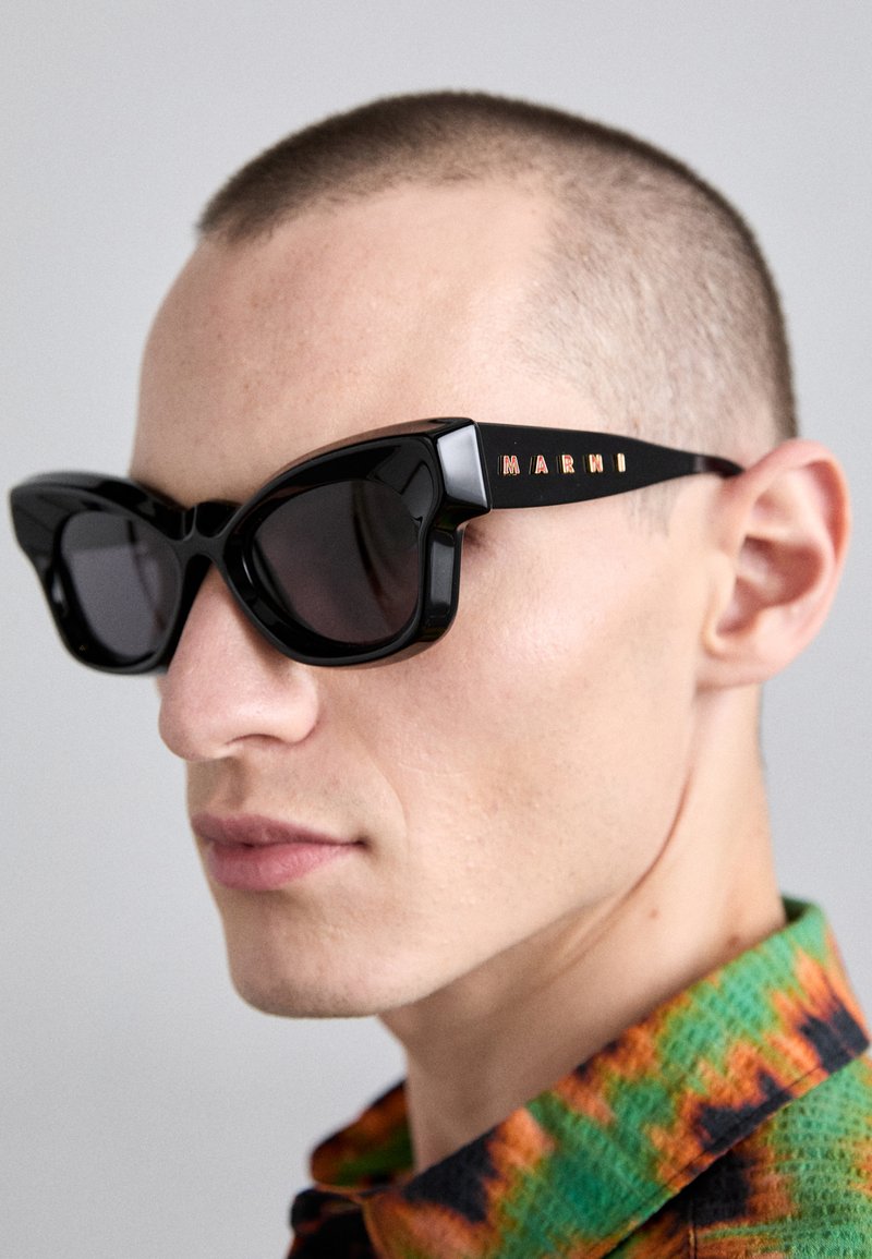 Occhiali da sole cat-eye neri con lenti scure, caratterizzati da una montatura spessa e il logo "MARNI" in lettere dorate sul lato, su uno sfondo chiaro.