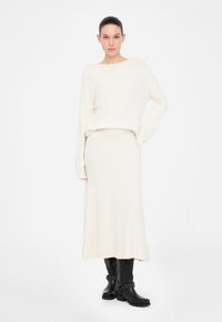 Suéter de punto color crema con escote amplio, combinado con una falda larga a juego. La modelo lleva botas de tacón negras con detalles en plateado.