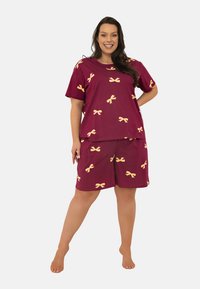 Uśmiechnięta kobieta stoi boso w bordowej bluzce z krótkim rękawem i dopasowanych szortach z żółtymi wzorami w kokardki na białym tle.