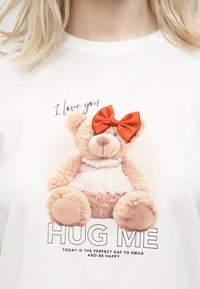 Αρκουδάκι με περιδέραιο από πέρλες και κόκκινο φιόγκο σε λευκό πουκάμισο, συνοδευόμενο από το κείμενο «Σ' αγαπώ», «ΑΓΚΑΛΙΑΣΕ ΜΕ» και ένα θετικό μήνυμα από κάτω.