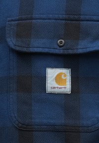 Μπλε και μαύρη καρό φανελένια μπλούζα με μπροστινή τσέπη, διαθέτοντας ραμμένο λογότυπο Carhartt και μαύρη λεπτομέρεια κουμπιού.