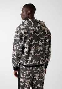 Felpa con cappuccio grigio scuro con stampa camouflage astratta color crema. Presenta polsini e orlo elasticizzati, progettata per comfort e libertà di movimento.