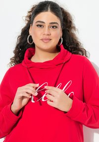 Punainen hupullinen collegepaita, jossa on valkoinen "Hello" -painatus. Kangas näyttää pehmeältä, ja siinä on rento malli sekä narut kaula-aukossa.