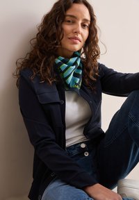 Mønstret multicolor tørklæde i blå og grønne nuancer, båret med en mørk jakke og hvid skjorte, parret med blå jeans og hvide sko.