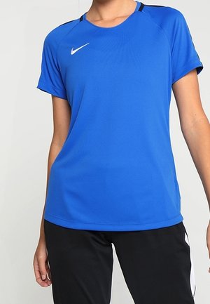 Personne portant un maillot de sport bleu à manches courtes avec un logo Nike blanc sur la poitrine et un pantalon de sport noir avec des bandes blanches sur les côtés.
