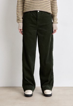 Person iført mørkegrønne kordfløjls-bredbenede bukser, cremefarvede loafers og en stribet beige og lysebrun langærmet top, stående mod en ensfarvet baggrund.
