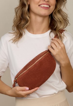 Õlgadeni blondidega naine naeratab, kandmas valget T-särki ja beežit seelikut, hoides roostekarva seemisnahast õlakotti kuldse lukkudega.