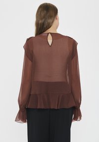 Sheer, rust-colored blouse with long sleeves, ruffled hem, and a keyhole detail at the back. Lightweight fabric with a textured finish.
