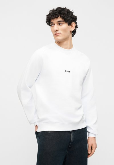 Jeune homme aux cheveux bouclés portant un sweat-shirt blanc MSGM et un jean foncé, regardant de côté sur un fond blanc uni.