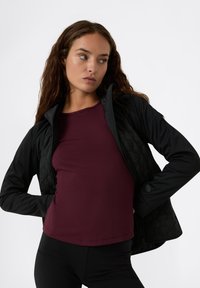 Mujer con cabello largo y rizado que lleva una chaqueta negra acolchada sobre una camiseta color marrón, posando con las manos en las caderas, mirando hacia un lado.