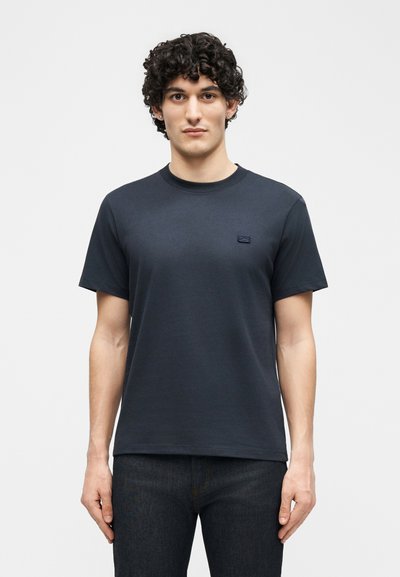 Νέος άνδρας με σγουρά μαλλιά φοράει ένα απλό σκούρο μπλε κοντομάνικο μπλουζάκι και σκούρα τζιν, στέκεται αντικρίζοντας μπροστά μπροστά σε ένα φωτεινό φόντο.