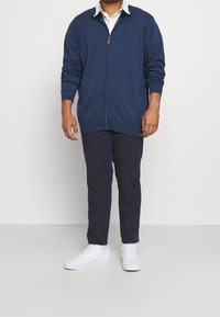 Námořnická modrá bunda na zip s dlouhými rukávy, pletená textura. Spárovaná s tmavými kalhotami a bílými teniskami. Jednoduchý design, žádné vzory ani akcenty.