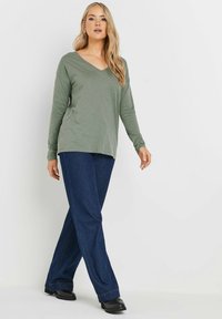 Langärmliges, V-Ausschnitt-Oberteil aus weichem olivgrünem Stoff, kombiniert mit dunkelblauen, weit geschnittenen Jeans. Schwarze Schuhe runden das Outfit ab. Insgesamt glatte Textur.