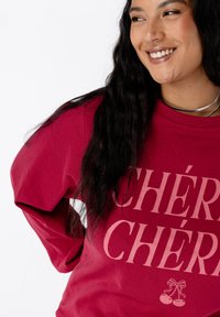 Personne souriante avec de longs cheveux noirs portant un sweat à capuche rouge avec le texte "CHÉRIE" et un graphique de cerise, des colliers en argent visibles.