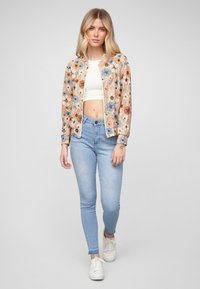 Veste bomber avec un motif floral dans des tons beiges, bleus et orange, associée à un jean skinny bleu clair et des baskets blanches.