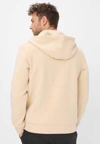 Felpa beige con cappuccio, dalla trama liscia, cuciture piatte e tasca a marsupio, mostrata da dietro su uno sfondo chiaro.