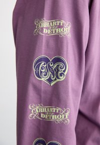Violetinė megzta maikė su auksiniu tekstu "Carhartt WIP Detroit" ir dekoratyvine širdimi, kurioje stilizuotu šriftu užrašyta "ONE". Lietimosi malonumo tekstūra, kasdieniškas dizainas.