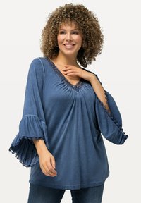 Blusa blu jeans con maniche a campana, scollatura a V e dettagli in pizzo floreale lungo il collo e i bordi delle maniche. Tessuto morbido e texturizzato.
