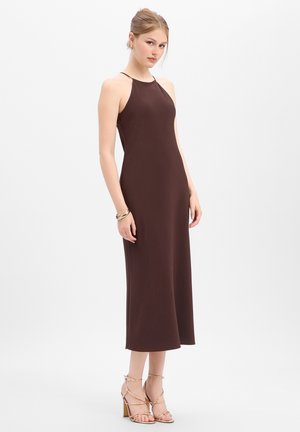 Young woman wearing a sleeveless brown midi dress and gold strappy high heels against a plain white background.