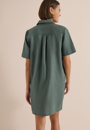 Femme aux cheveux courts bruns portant une robe-chemise verte fluide à manches courtes, debout de dos devant un fond beige uni.