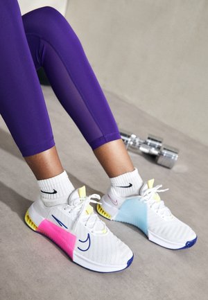 Piernas con leggings morados, calcetines blancos de Nike y zapatillas deportivas blancas y coloridas con acentos rosas y azules, descansando sobre un suelo gris cerca de mancuernas plateadas.