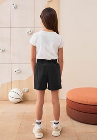 Svarta shorts med elastisk midja och en vit kortärmad skjorta. Bakgrunden har en klättervägg och ett runt orange sittområde.