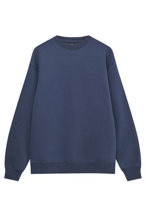 Mand med ryggen til iført en løs navy sweatshirt, brede cremefarvede bukser med baglommer og hvide sneakers på en hvid baggrund.
