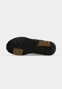 Semelle de chaussure noire avec un motif de bande de roulement rugueux et deux patches en caoutchouc marron pour la traction, photographiée sur un fond blanc uni.