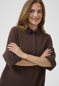 Ženska s svetlo rjavimi lasmi, oblečena v rjavo srajco z zavihanimi rokavi, prekrižanimi rokami, rahlo se smeji na enobarvnem ozadju.