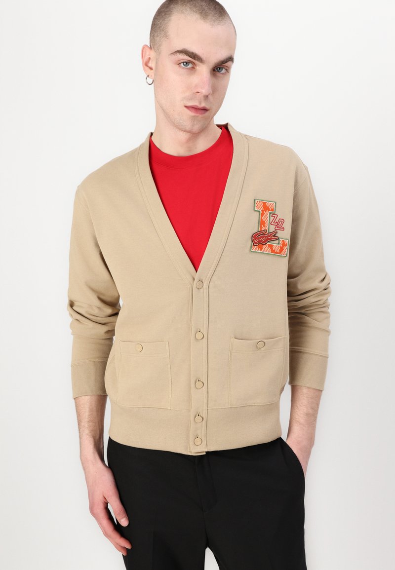 Cardigan beige avec un col en V, deux poches avant et fermeture par boutons. Il présente un patch brodé coloré sur le côté gauche.