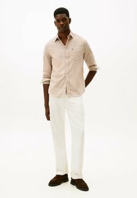 Beige button-up skjorte med en brystlomme, iført hvide bukser og brune sko, vist på en model mod en ensfarvet baggrund.