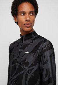 Schwarzes Sportoberteil mit hohem Kragen, ausgestattet mit einem Reißverschluss und strukturierten Grafikmustern in verschiedenen Schwarzschattierungen sowie einem kleinen weißen Logo.