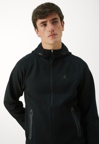 Schwarzer Kapuzenpullover mit durchgehendem Reißverschluss, zwei seitlichen Taschen mit eleganten Reißverschlüssen und einem kleinen Logo auf der Brust. Weiche Stofftextur.