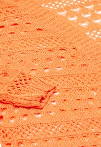 Orange stickat tyg med öppna mönster, featuring texturerade ränder och en liten näsdukliknande bit vikt vid kanten.