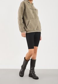 Fleece-hættetrøje i olivengrøn med kængurulomme og snøre i hætten, kombineret med sorte biker-shorts og knælange sorte støvler med spænde detaljer.