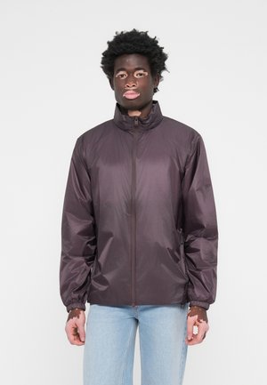 Purple windbreaker jacket with a high collar, full-length zipper, elastic cuffs, and a lightweight, shiny texture. Worn over light denim jeans.