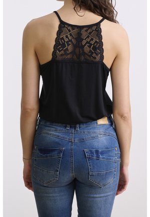 Kobieta w czarnej, bezrękawnikowej bluzce z koronkowym tyłem i niebieskich dżinsach, widziana od tyłu na jednolitym tle.