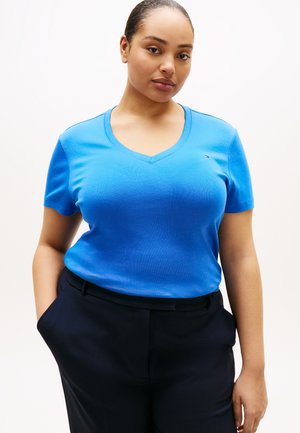 Woman wearing a bright blue V-neck short-sleeve shirt tucked into black pants, posing with hands in pockets against a plain background.